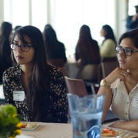 Two young women wearing glasses sit at a table and listen to the conversation at the table.