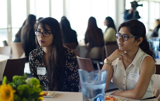 Two young women wearing glasses sit at a table and listen to the conversation at the table.