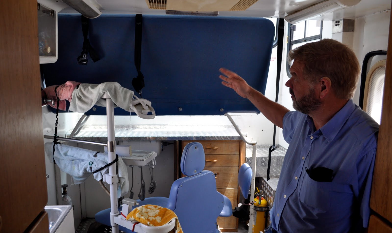Man standing inside a trailer with dental chair and equipment, gesturing toward the equipment