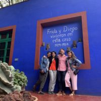 Four people pose for photo in front of blue structure. Text on wall says Frida y Diego vivieron en esta casa.