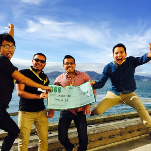 four people holding a large check. the person on the right end is jumping excitedly with both feet in the air